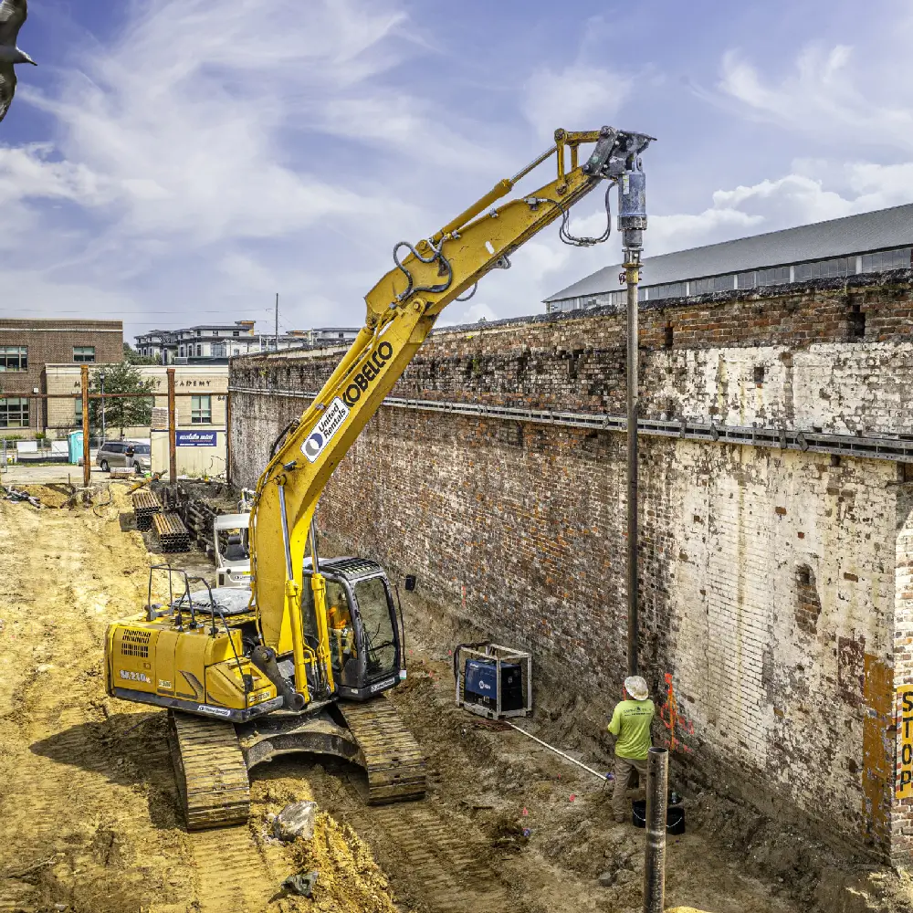 Excavator at construction site with worker and old brick wall.